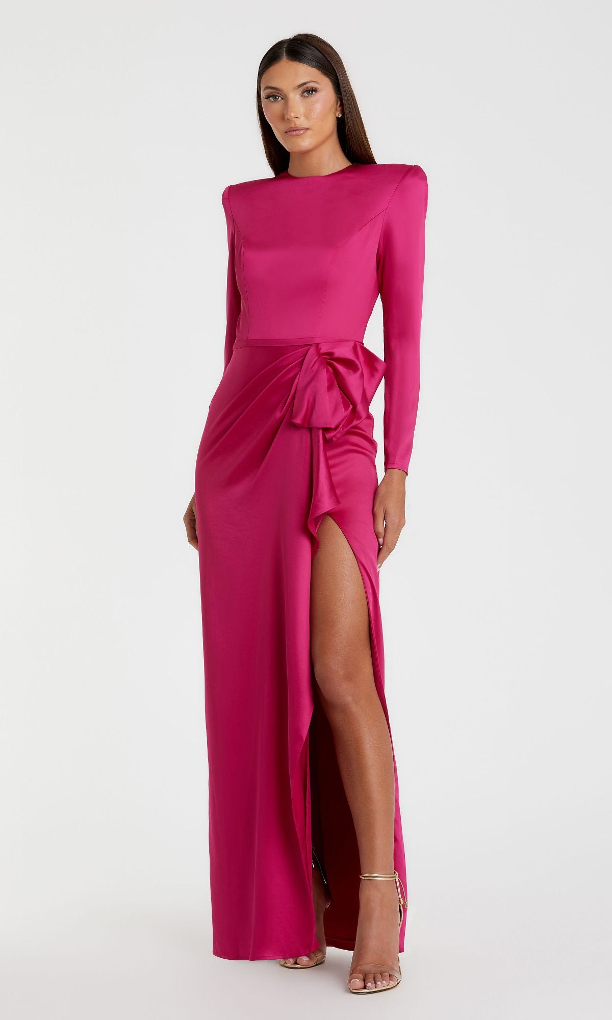 A woman models the Mac Duggal 26676 Bold-Shoulder Long Formal Dress, a vibrant pink, bra-friendly evening gown with long sleeves, a side slit, and a waist bow, posed against a simple white background.